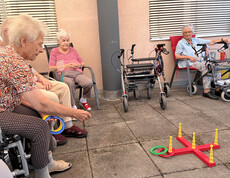 Bewohner*innen sitzen im Kreis und nehmen an einem Ringwurfspiel auf der Terrasse teil.
