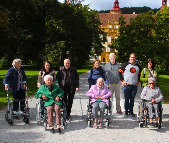 Drei Bewohnerinnen sitzen in Rollstühlen nebeneinander, eine weitere Bewohnerin steht mit einem Rollator etwas seitlich. Hinter ihnen befinden sich Pflegepersonal sowie Angehörige. Im Hintergrund ist das Schloss Eggenberg zu sehen.