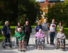 Drei Bewohnerinnen sitzen in Rollstühlen nebeneinander, eine weitere Bewohnerin steht mit einem Rollator etwas seitlich. Hinter ihnen befinden sich Pflegepersonal sowie Angehörige. Im Hintergrund ist das Schloss Eggenberg zu sehen.