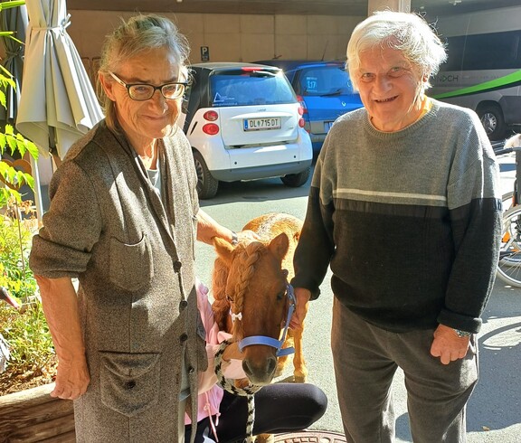 Zwei Bewohnerinnen stehen neben dem kleinen Pony Maxi und streicheln es, während eine Begleitperson das Pony im Außenbereich des Pflegewohnhauses hält.
