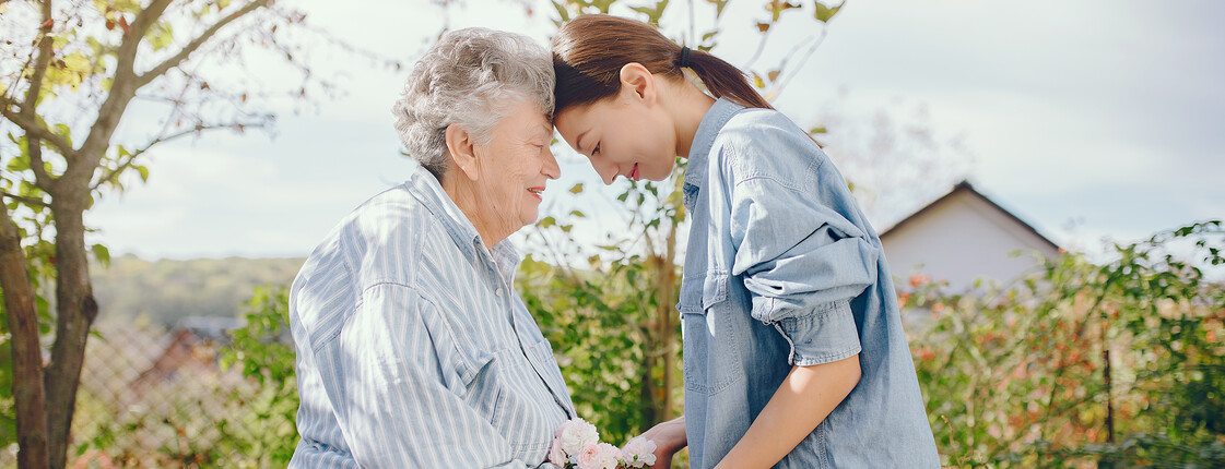 Eine ältere Frau mit grauen Haaren und eine junge Frau stehen in einem Garten, lehnen ihre Stirn aneinander und halten gemeinsam einen kleinen Blumenstrauß.