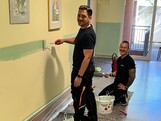 Zwei Männer – einer steht, der andere kniet – lächeln im Gang eines Pflegewohnhauses und blicken direkt in die Kamera. Beide halten eine Farbrolle in der Hand. Sie sind dabei, den unteren Wandbereich in einem freundlichen Mintgrün zu streichen. Die Atmosphäre wirkt fröhlich und engagiert.