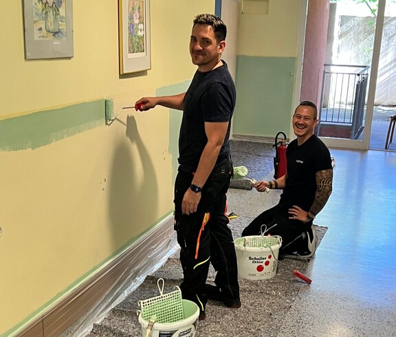 Zwei Männer – einer steht, der andere kniet – lächeln im Gang eines Pflegewohnhauses und blicken direkt in die Kamera. Beide halten eine Farbrolle in der Hand. Sie sind dabei, den unteren Wandbereich in einem freundlichen Mintgrün zu streichen. Die Atmosphäre wirkt fröhlich und engagiert.