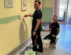 Zwei Männer – einer steht, der andere kniet – lächeln im Gang eines Pflegewohnhauses und blicken direkt in die Kamera. Beide halten eine Farbrolle in der Hand. Sie sind dabei, den unteren Wandbereich in einem freundlichen Mintgrün zu streichen. Die Atmosphäre wirkt fröhlich und engagiert.