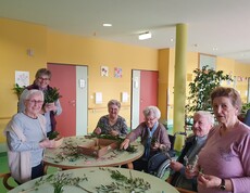 Bewohnerinnen und Bewohner des Pflegewohnhauses sitzen gemeinsam an mehreren Tischen und binden Palmbuschen. Zwischen frischen Zweigen, Bändern und Bastelmaterial entstehen in gemütlicher Runde kunstvolle Ostersträuße. Die fröhliche Aktivität findet im farbenfrohen Aufenthaltsbereich statt.