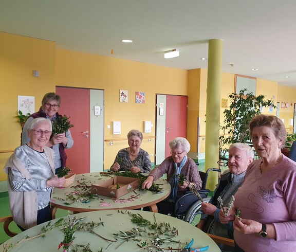 Bewohnerinnen und Bewohner des Pflegewohnhauses sitzen gemeinsam an mehreren Tischen und binden Palmbuschen. Zwischen frischen Zweigen, Bändern und Bastelmaterial entstehen in gemütlicher Runde kunstvolle Ostersträuße. Die fröhliche Aktivität findet im farbenfrohen Aufenthaltsbereich statt.