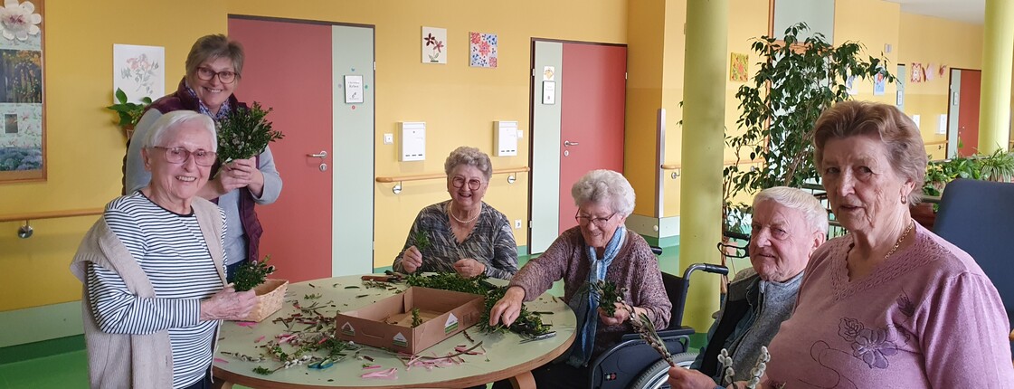 Bewohnerinnen und Bewohner des Pflegewohnhauses sitzen gemeinsam an mehreren Tischen und binden Palmbuschen. Zwischen frischen Zweigen, Bändern und Bastelmaterial entstehen in gemütlicher Runde kunstvolle Ostersträuße. Die fröhliche Aktivität findet im farbenfrohen Aufenthaltsbereich statt.