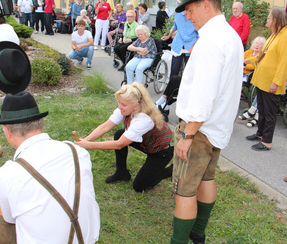 Mehrere Mitglieder der Pischelsdorfer Schuhplattler in Tracht demonstrieren das traditionelle Umlegen des Maibaums, während Bewohnerinnen und Bewohner des Caritas Pflegewohnhauses das Geschehen aus nächster Nähe verfolgen.