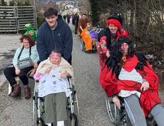Verkleidete Bewohner*innen werden im Freien im Rollstuhl begleitet, während die Gruppe gemeinsam spaziert.