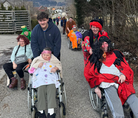 Verkleidete Bewohner*innen werden im Freien im Rollstuhl begleitet, während die Gruppe gemeinsam spaziert.
