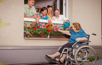 Vier Pflegekräfte lehnen sich aus dem Fenster, eine Bewohnerin sitzt im Rollstuhl und gießt die Blumen die auf der Fensterbank stehen.
