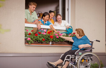 Vier Pflegekräfte lehnen sich aus dem Fenster, eine Bewohnerin sitzt im Rollstuhl und gießt die Blumen die auf der Fensterbank stehen.