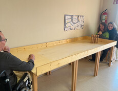 Bewohner stößt eine Kugel über die Holzbahn, zwei Pflegekräfte stehen am anderen Ende und beobachten das Geschehen.