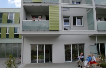 Ansicht des Hauses, auf einem Balkon im ersten Stock stehen zwei Frauen uns sehen auf zwei Frauen uns einen Mann herunter, die vor dem Haus auf einer Bank sitzen. 