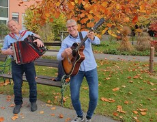 Vor dem Pflegewohnhaus stehen zwei Personen unter einem Baum mit leuchtend orangefarbenem Herbstlaub. Sie musizieren gemeinsam – eine Person spielt Gitarre, die andere Akkordeon. Im Vordergrund liegen herabgefallene Blätter, im Hintergrund sind Wege und herbstlich dekorierte Grünflächen zu sehen. Die Szene strahlt herbstliche Stimmung und musikalische Geselligkeit aus.