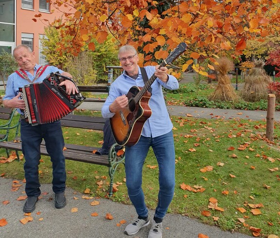 Vor dem Pflegewohnhaus stehen zwei Personen unter einem Baum mit leuchtend orangefarbenem Herbstlaub. Sie musizieren gemeinsam – eine Person spielt Gitarre, die andere Akkordeon. Im Vordergrund liegen herabgefallene Blätter, im Hintergrund sind Wege und herbstlich dekorierte Grünflächen zu sehen. Die Szene strahlt herbstliche Stimmung und musikalische Geselligkeit aus.
