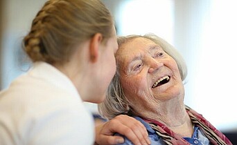 Ältere Damen sitzt und lacht, eine junge Frau sitzt neben ihr und berührt ihre Schulter