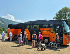 Senior*innen steigen mit Rollstühlen aus einem orangefarbenen Reisebus im Grünen.