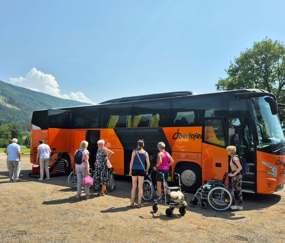 Senior*innen steigen mit Rollstühlen aus einem orangefarbenen Reisebus im Grünen.