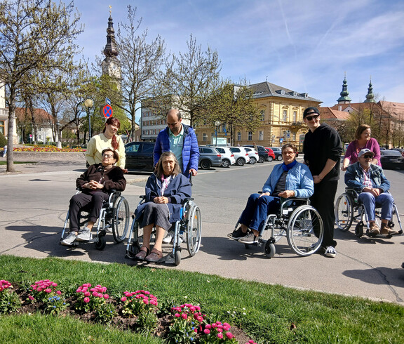 Gruppe von Menschen im Rollstuhl und Begleitpersonen macht Halt an einem Platz mit Frühlingsblumen in Klagenfurt.