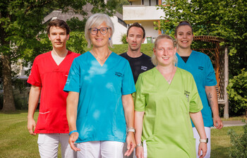 Mitarbeiter*innen in bunten Dienstkleidungen stehen im Garten eines Pflegewohnhauses und lächeln in die Kamera.