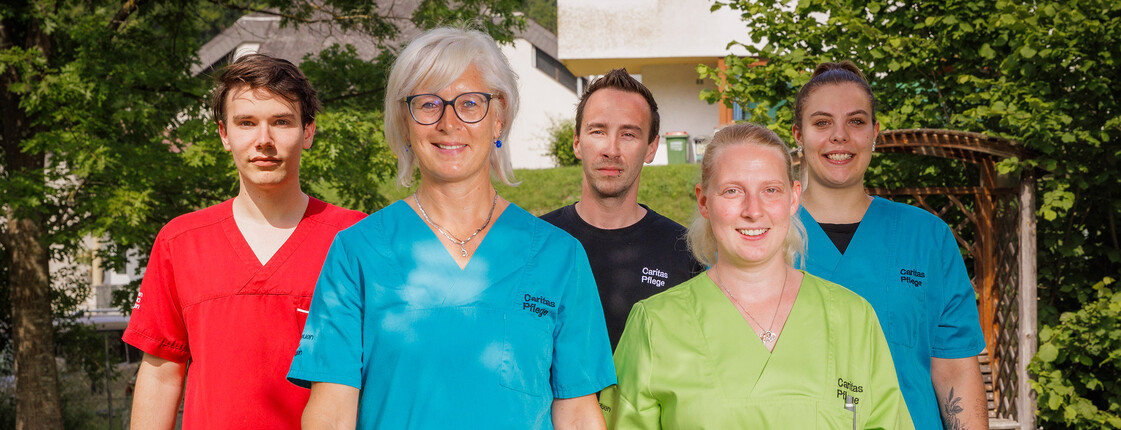 Mitarbeiter*innen in bunten Dienstkleidungen stehen im Garten eines Pflegewohnhauses und lächeln in die Kamera.