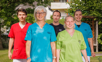 Mitarbeiter*innen in bunten Dienstkleidungen stehen im Garten eines Pflegewohnhauses und lächeln in die Kamera.