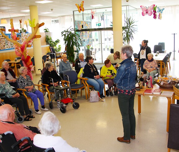 Im liebevoll dekorierten Gemeinschaftsbereich des Pflegewohnhauses sitzen zahlreiche Bewohnerinnen und Bewohner in einer großen Runde beisammen. Wortgottesdienst Leiterin Eva Kaspar steht vor der Gruppe und spricht zu ihnen, während auf einem Tisch dekorative Palmbuschen und Geschenke bereitstehen.