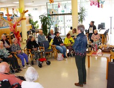 Im liebevoll dekorierten Gemeinschaftsbereich des Pflegewohnhauses sitzen zahlreiche Bewohnerinnen und Bewohner in einer großen Runde beisammen. Wortgottesdienst Leiterin Eva Kaspar steht vor der Gruppe und spricht zu ihnen, während auf einem Tisch dekorative Palmbuschen und Geschenke bereitstehen.
