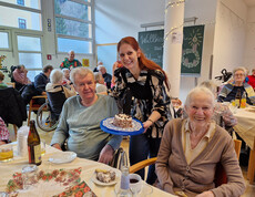 Person präsentiert Kuchen an zwei Gäste, festlich gedeckter Tisch im Pflegewohnhaus