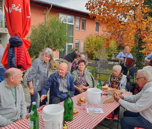 Im Garten vor dem Pflegewohnhaus sitzen Bewohnerinnen und Bewohner an mehreren Tischen und genießen frisch gebratene Maroni. Die herbstliche Stimmung wird durch das gemütliche Beisammensein und die musikalische Begleitung im Hintergrund, wo die Musikanten zu sehen sind, wunderbar unterstrichen.