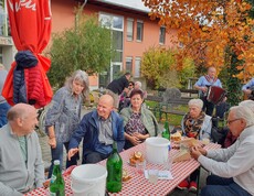 Im Garten vor dem Pflegewohnhaus sitzen Bewohnerinnen und Bewohner an mehreren Tischen und genießen frisch gebratene Maroni. Die herbstliche Stimmung wird durch das gemütliche Beisammensein und die musikalische Begleitung im Hintergrund, wo die Musikanten zu sehen sind, wunderbar unterstrichen.
