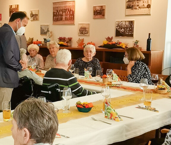 Eine gemütliche Runde beim Erntedankfest. Ältere Menschen sitzen an einem herbstlich dekorierten Tisch, während ein Mann mit ihnen spricht. Der Raum ist festlich geschmückt, passend zum Anlass des Erntedankfestes.