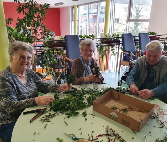 Mehrere Bewohnerinnen und Bewohner des Pflegewohnhauses sitzen gemeinsam am Tisch und binden Palmbuschen. Vor ihnen liegen frische Zweige, Bänder und Werkzeuge, während in der hellen, freundlichen Umgebung konzentriert gearbeitet und miteinander gesprochen wird.