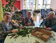 Mehrere Bewohnerinnen und Bewohner des Pflegewohnhauses sitzen gemeinsam am Tisch und binden Palmbuschen. Vor ihnen liegen frische Zweige, Bänder und Werkzeuge, während in der hellen, freundlichen Umgebung konzentriert gearbeitet und miteinander gesprochen wird.