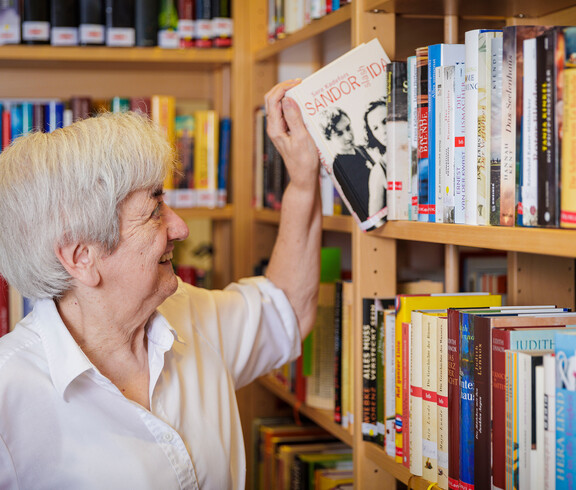 Eine Dame lächelt und zieht ein Buch aus einem Bücherregal.