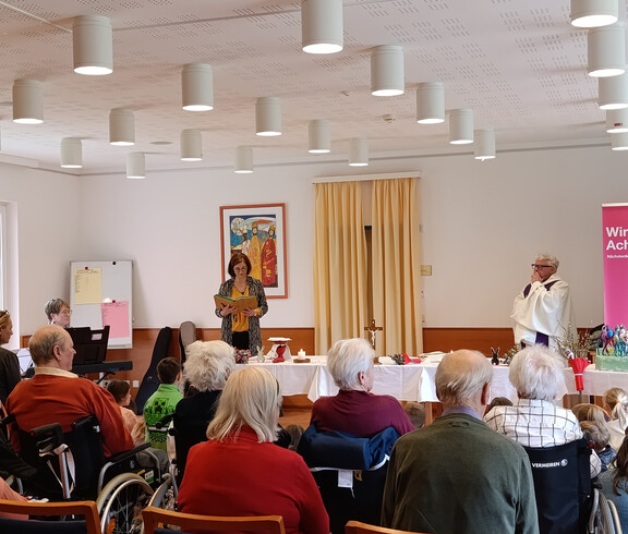 Frau steht vor einer Gruppe von Bewohner*innen und Kindern und liest etwas ab.
