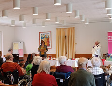 Frau steht vor einer Gruppe von Bewohner*innen und Kindern und liest etwas ab.