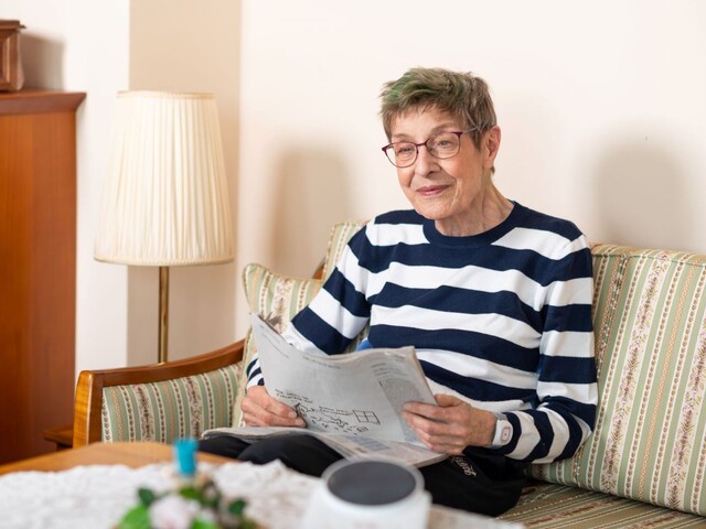 Ältere Frau mit grau-grünen Haaren und Brille sitzt lächelnd auf dem Sofa und liest Zeitung, im Vordergrund ein Telefonhörer-Symbol als Verweis auf das Caritas Notruftelefon