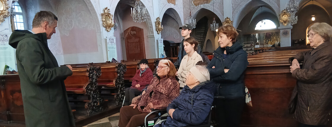 Gruppe älterer Menschen, einige in Rollstühlen, hören dem Pfarrer in einer Kirche zu.