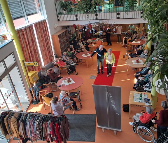 Von oben ist eine Modeschau in einem barrierefreien Gemeinschaftsbereich des Pflegewohnhauses zu sehen, mit rotem Laufsteg in der Mitte und sitzendem Publikum, teils mit Rollstühlen und Gehhilfen.