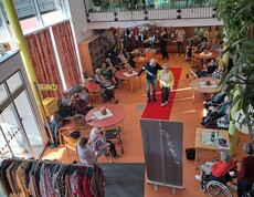 Von oben ist eine Modeschau in einem barrierefreien Gemeinschaftsbereich des Pflegewohnhauses zu sehen, mit rotem Laufsteg in der Mitte und sitzendem Publikum, teils mit Rollstühlen und Gehhilfen.