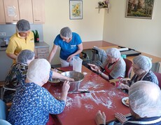 Bewohnerinnen und Bewohner des Pflegewohnhauses sitzen gemeinsam am großen Tisch und bereiten Germteig für österliche Backwaren zu. Betreuungskräfte unterstützen beim Kneten, Formen und Portionieren, während Mehl, Teig und Backutensilien für eine lebendige, fröhliche Backatmosphäre sorgen.