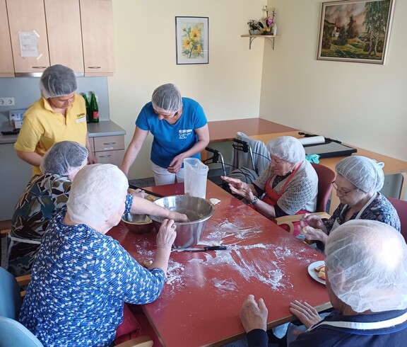 Bewohnerinnen und Bewohner des Pflegewohnhauses sitzen gemeinsam am großen Tisch und bereiten Germteig für österliche Backwaren zu. Betreuungskräfte unterstützen beim Kneten, Formen und Portionieren, während Mehl, Teig und Backutensilien für eine lebendige, fröhliche Backatmosphäre sorgen.