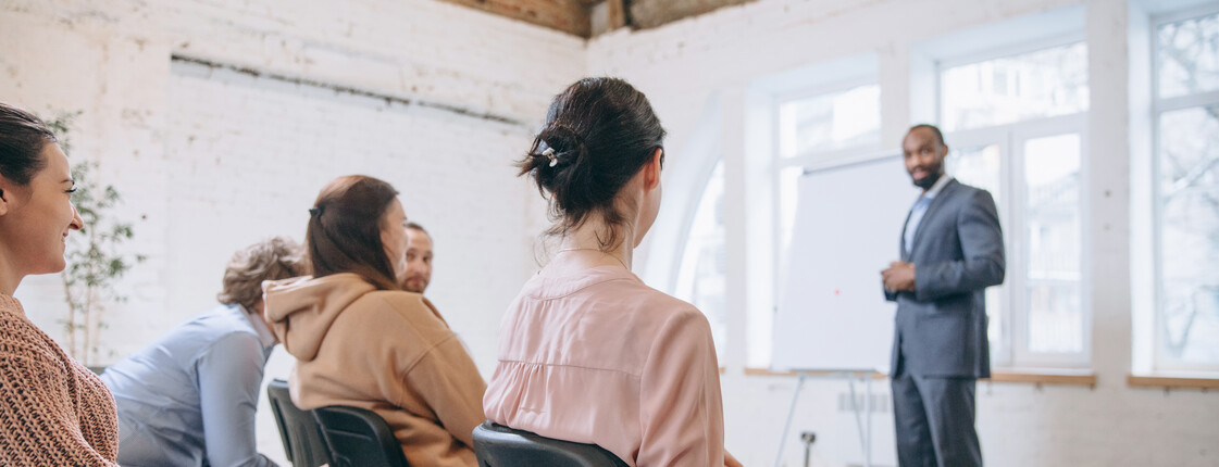 Inhouse-Schulungen für Unternehmen Ein Vortragender gibt bei einem universitären Workshop einen Vortrag. Die Zuhörenden in der ersten Reihe des Publikums sind unkenntlich.