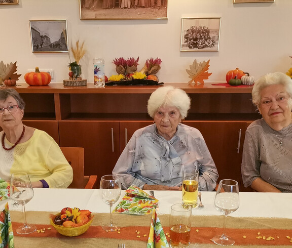 Drei Damen, die an einem festlich gedeckten Tisch sitzen. Der Tisch ist herbstlich dekoriert mit Erntedank-Motiven wie Kürbissen und Blumen. Die Atmosphäre wirkt herzlich und einladend.