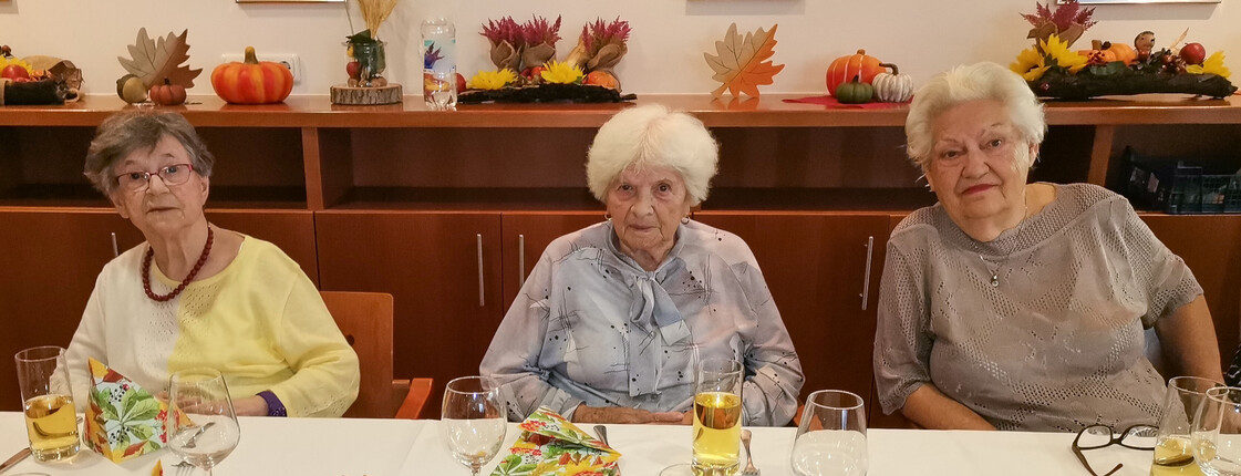 Drei Damen, die an einem festlich gedeckten Tisch sitzen. Der Tisch ist herbstlich dekoriert mit Erntedank-Motiven wie Kürbissen und Blumen. Die Atmosphäre wirkt herzlich und einladend.