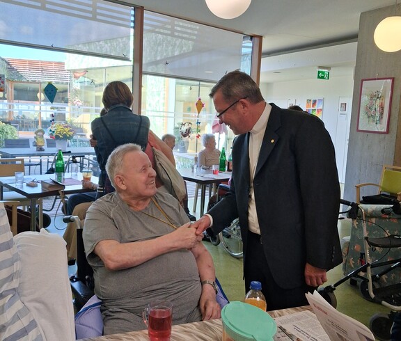 Ein Bewohner sitzt im Aufenthaltsraum des Caritas Pflegewohnhauses, während Weihbischof Johannes Freitag die Person mit einem Handschlag begrüßt und sich im persönlichen Gespräch austauscht.