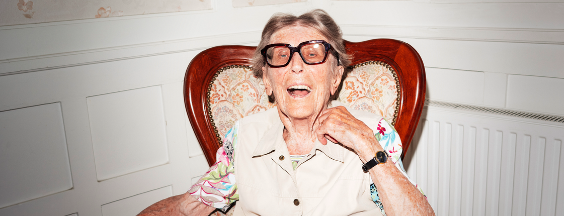 Es ist das Porträt einer älteren Dame zusehen, sie sitzt in einem Ohrensessel und lacht in die Kamera.