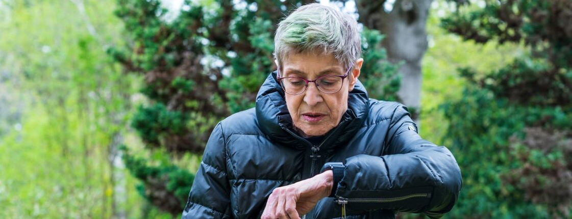 Person im Freien trägt eine dunkle Jacke und blickt auf ein Armband mit integriertem Caritas-Notrufsystem am Handgelenk; im Hintergrund sind Bäume und Grünflächen zu sehen.